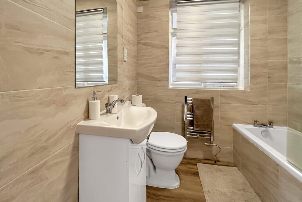 A tiled family bathroom with a bath, separate shower attachment, pedestal basin, WC, chrome heated towel rail, and two frosted windows.