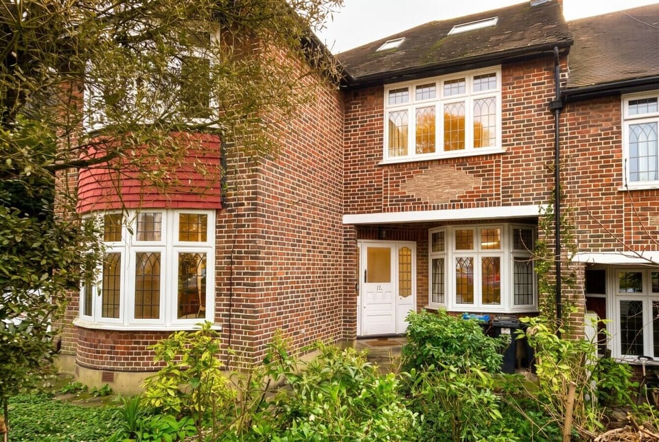 The exterior of the property showing a red-brick semi-detached house with leaded bay windows, a white front door, and mature front garden planting.