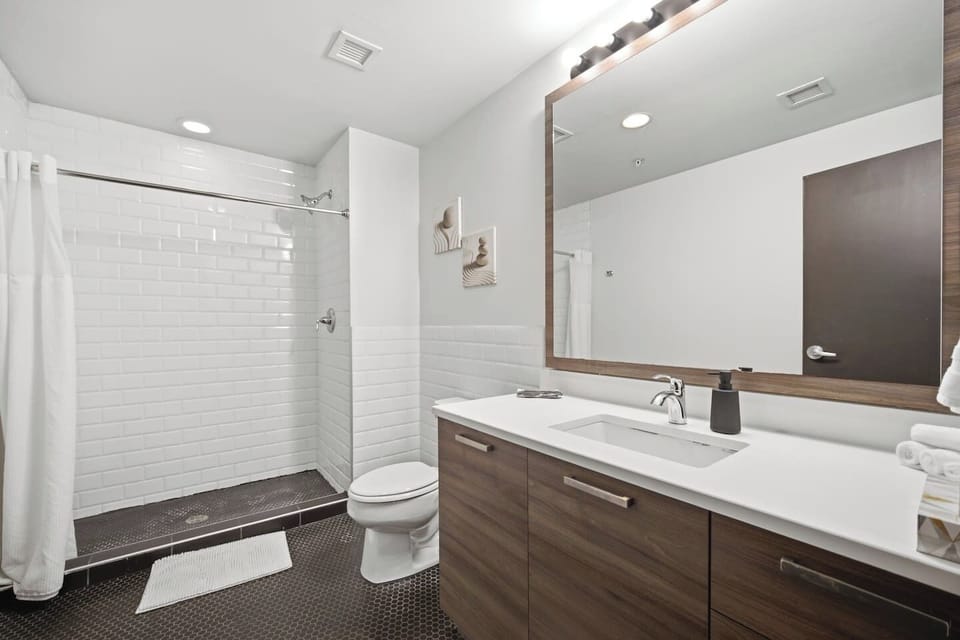 Stylish apartment bathroom featuring a vanity with a white countertop, a large mirror, a tiled shower stall with a white curtain, and white subway tile accents.