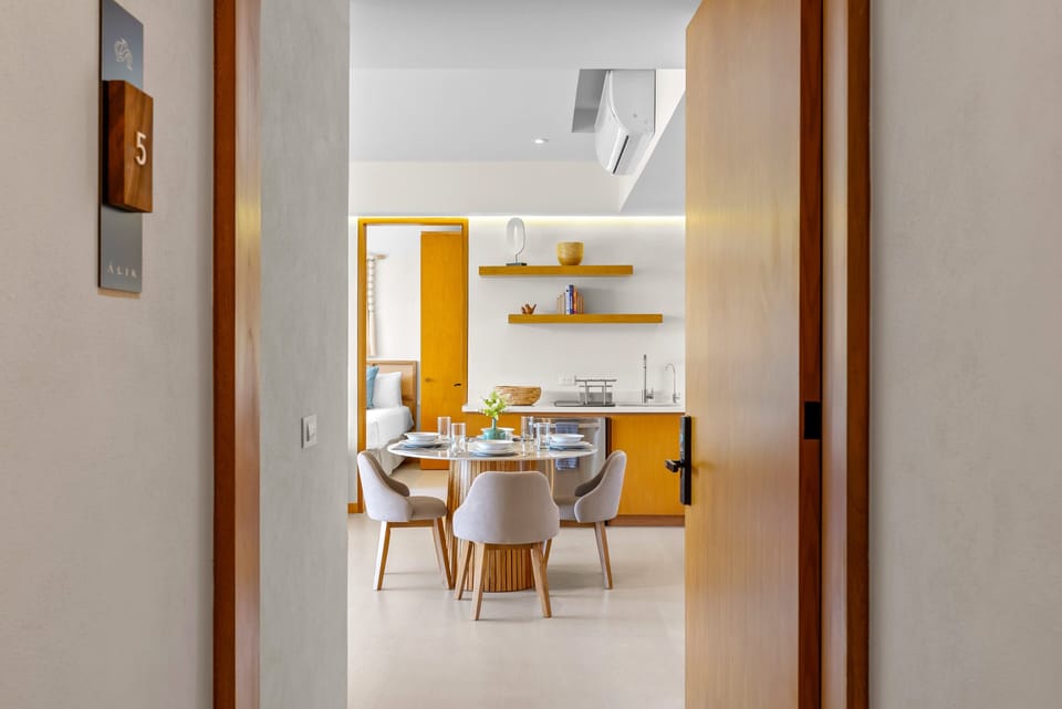 Open-door perspective from unit 5 hallway entering into an open-concept dining area, modern wooden kitchen with floating shelves, and a glimpse beyond into the sunlit bedroom suite.