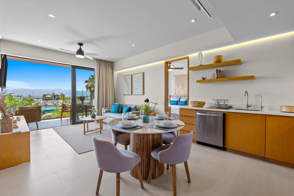 Sunlit, open-concept suite combining dining, living, and kitchen areas, flowing seamlessly onto a private ocean-view balcony overlooking the marina through large floor-to-ceiling glass doors.