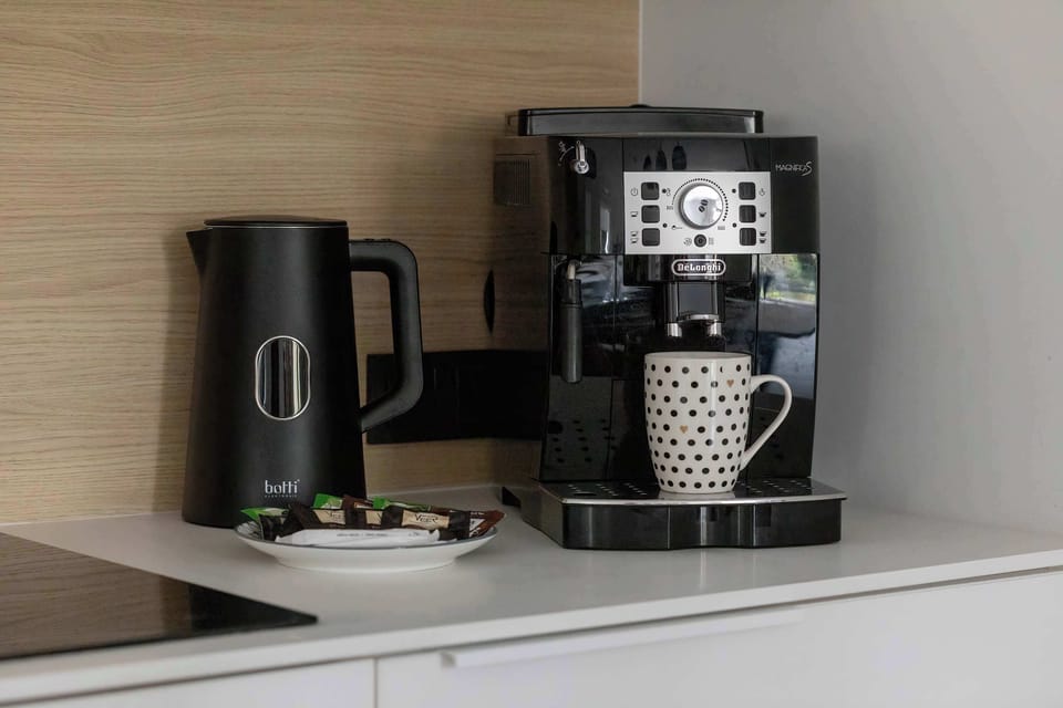 A close-up shot displays a black coffee machine and electric kettle resting on the kitchen counter next to a mug on a small saucer.