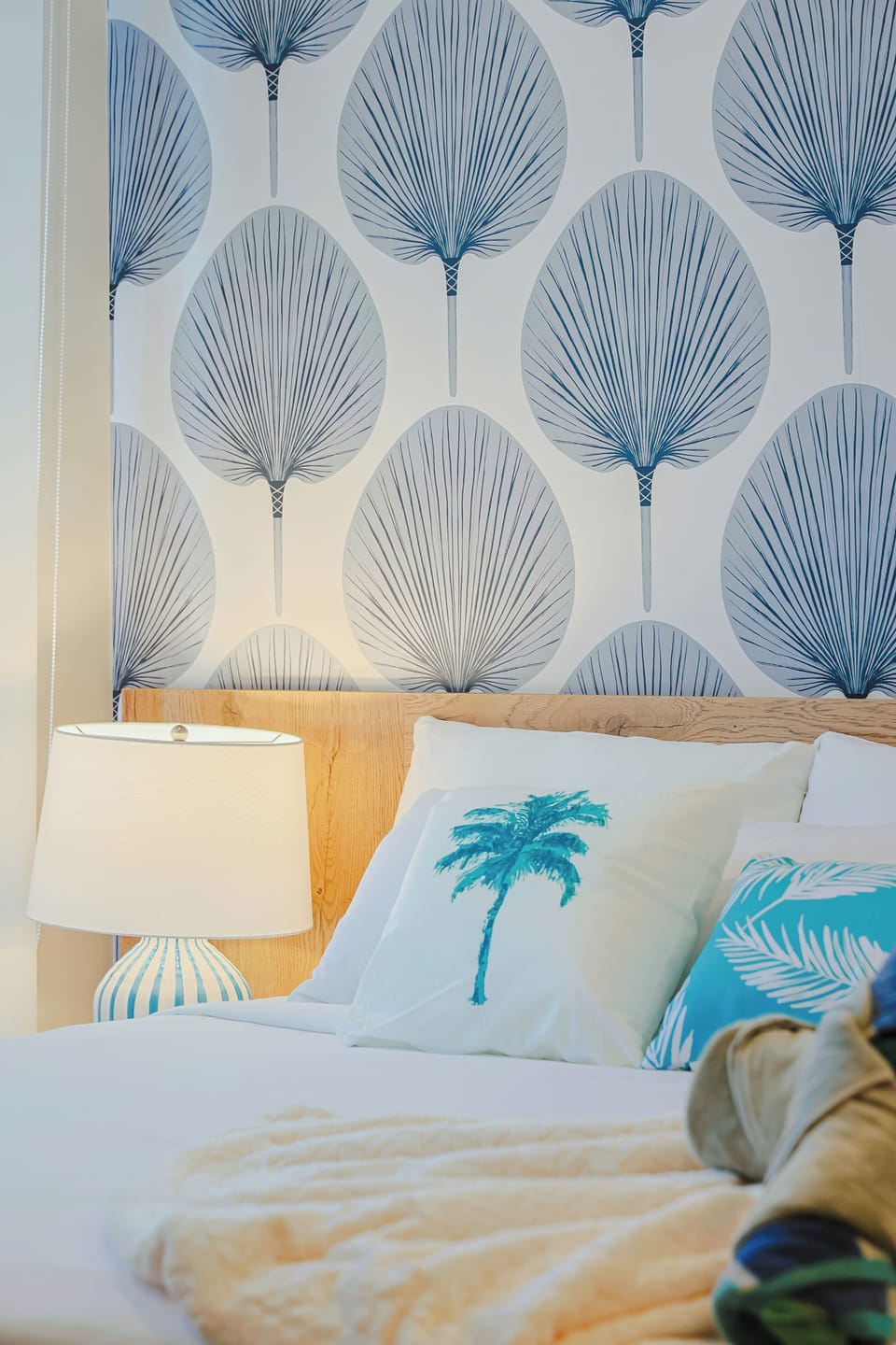 A cozy and refreshing bedroom accented by soft blue tones, tropical-inspired pillows, and a beautiful botanical wall design. Crisp bedding, warm wood details, and gentle lighting create a serene and inviting space perfect for relaxing after a day in 