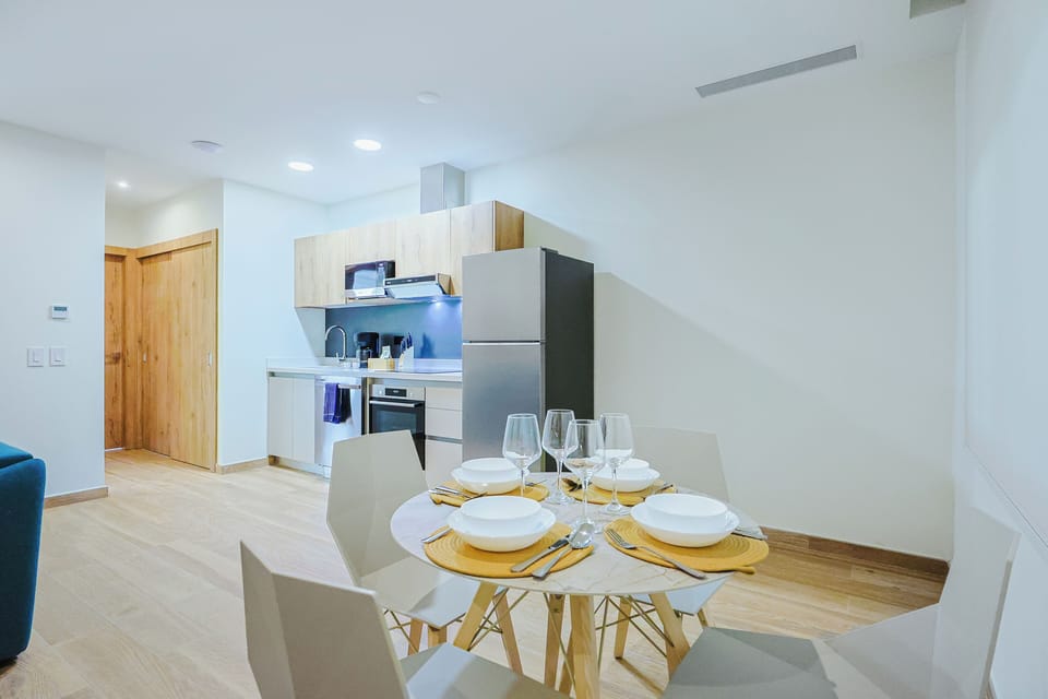 A bright, modern open-plan kitchen and dining space, elegantly set for four with golden placemats and fresh glassware. The sleek compact kitchen offers everything you need to prepare meals during your stay.