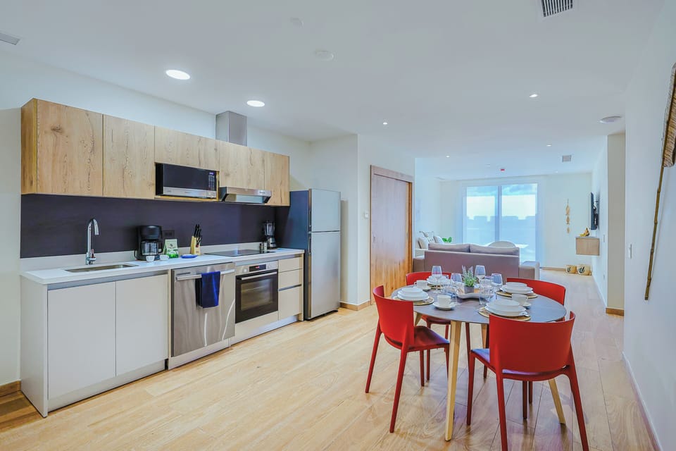Your bright, open kitchen flows into a cheerful dining area with bold red chairs, perfect for sharing meals. Through the doorway, a cozy living space awaits with natural light streaming in.