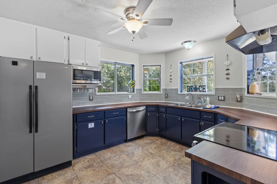 Cook up fresh catch in this bright, coastal kitchen with navy cabinets, wood counters, and stainless appliances, all framed by sunny garden windows.