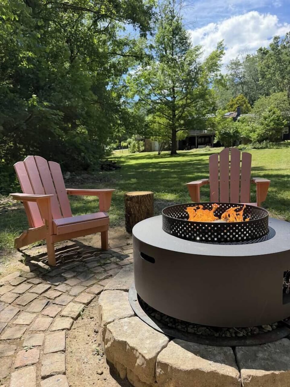 Outdoor fire pit with Adirondack chairs, surrounded by trees and a spacious green yard.