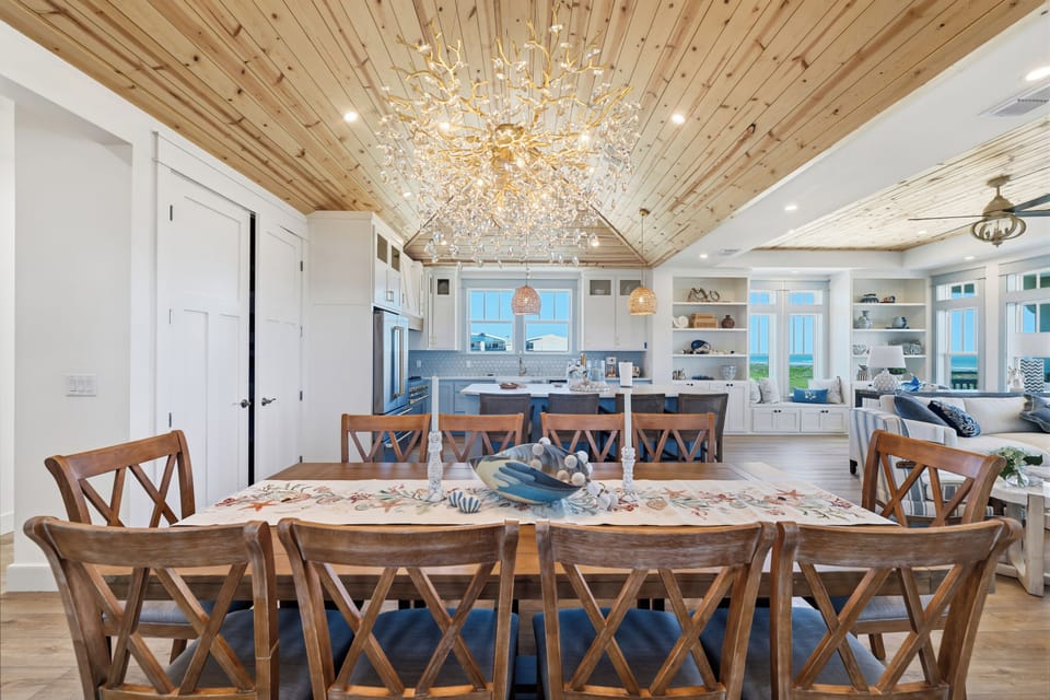 Gather around the stunning wood dining table beneath a sparkling chandelier and natural wood ceiling. The open layout flows seamlessly into your coastal kitchen and living area, where blue water views await through every window.