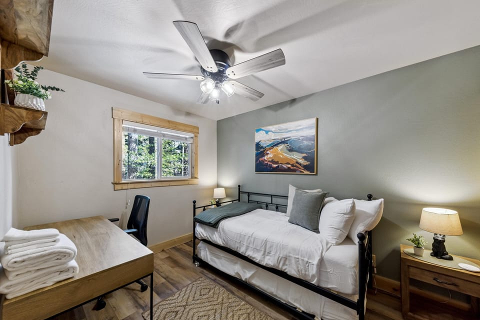 Single beds - trundle beds with white linens and gray pillows sits beneath a landscape painting, next to a wooden desk and window.