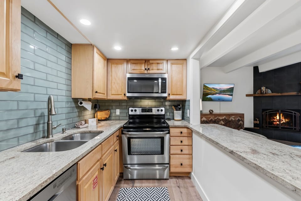 A welcoming kitchen awaits, with warm wood cabinetry, gleaming granite counters, and a cozy fireplace crackling nearby—perfect for preparing your favorite meals in comfort.