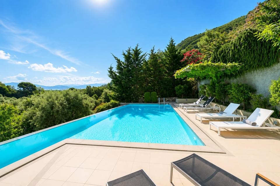 A sun-drenched outdoor swimming pool features lounge chairs arranged on a tiled patio, surrounded by dense green trees and a sweeping view of distant mountains.