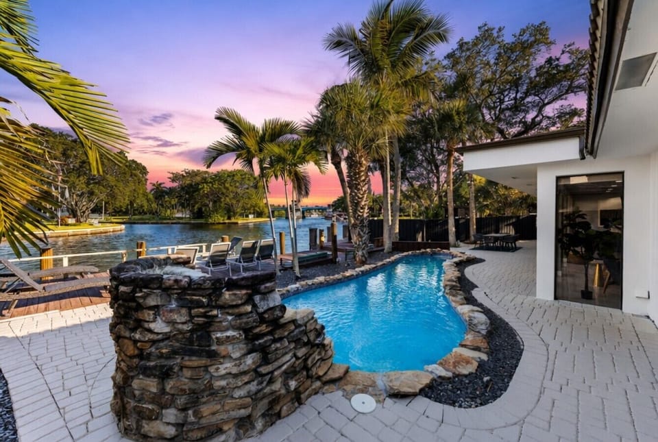 Custom pool deck with stacked-stone water feature, paver patio, palm trees, and wide-open views of the waterway at twilight