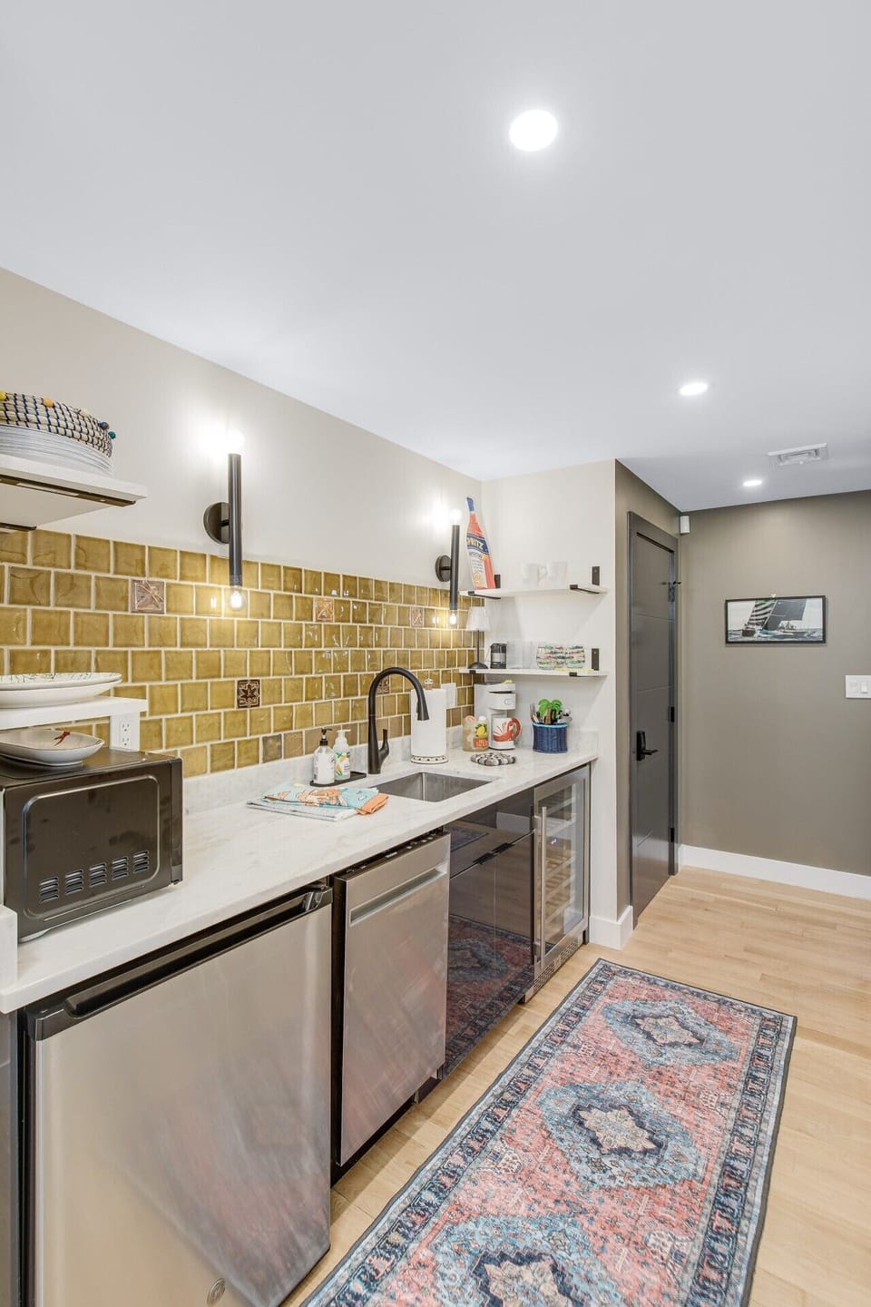 Functional kitchenette counter featuring an undermount sink with a black gooseneck faucet, olive-green tile backsplash, microwave, and open shelving.