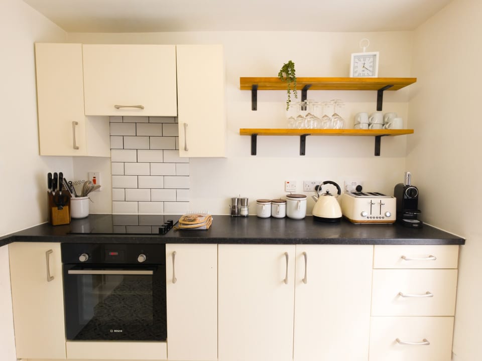 Cream units and a dark worktop, with open shelves for glasses and mugs. Oven, kettle and coffee machine all to hand.