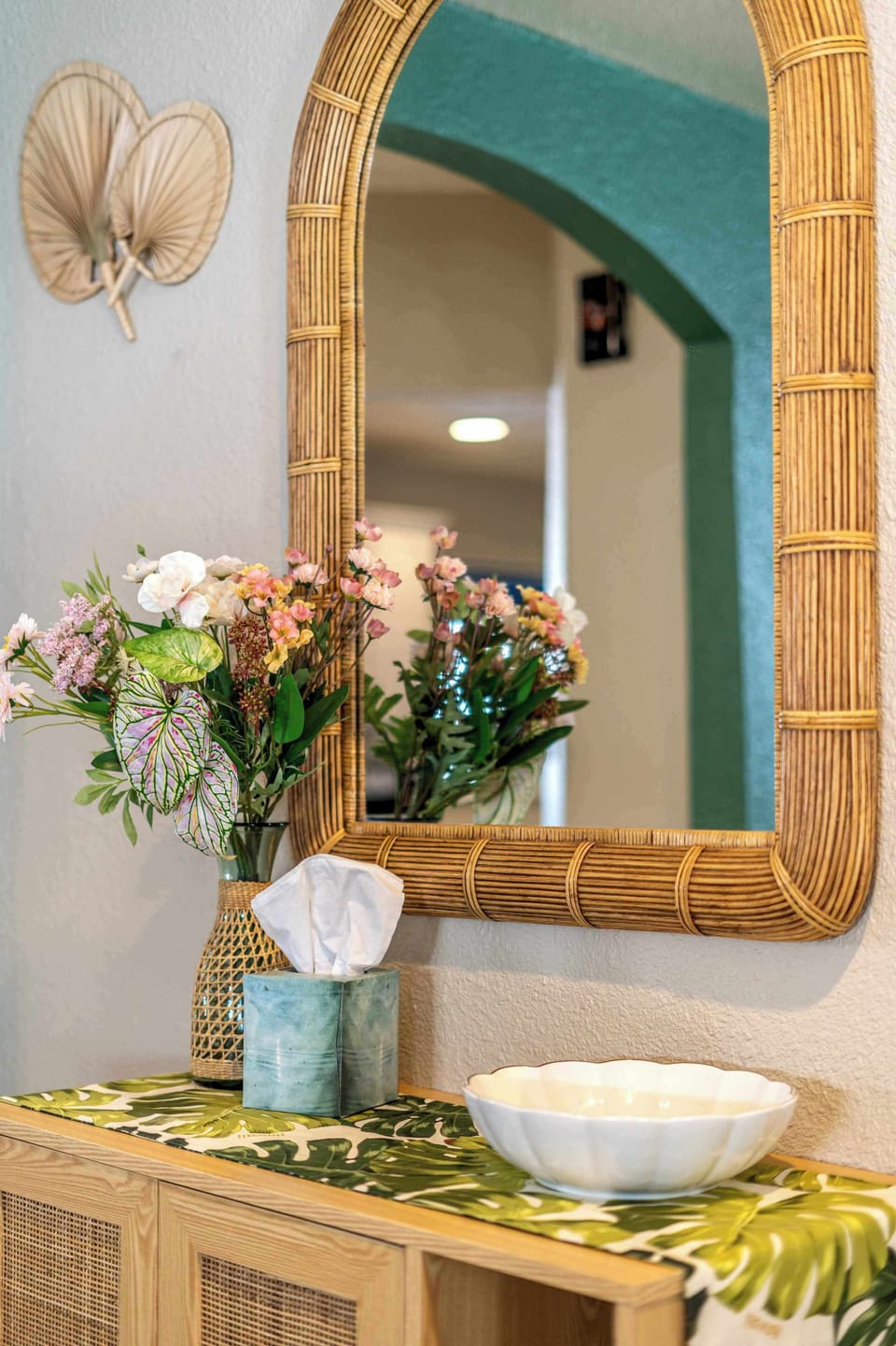 A rattan framed arch mirror hangs above a light wood console styled with fresh florals and a woven tray. The textured teal wall and brass starburst above give the entry a collected, lived in feel rather than a staged one