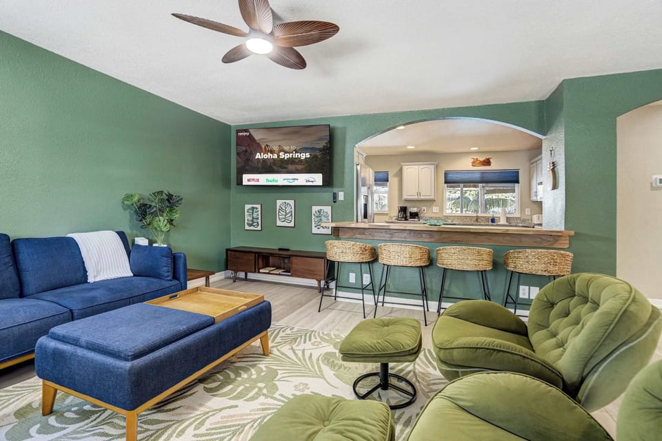 A wall mounted Smart TV hangs beside the arch, with the rustic bar and its three rattan stools bridging the living room and kitchen. Drinks get poured here before dinner, close enough that nobody misses the conversation.