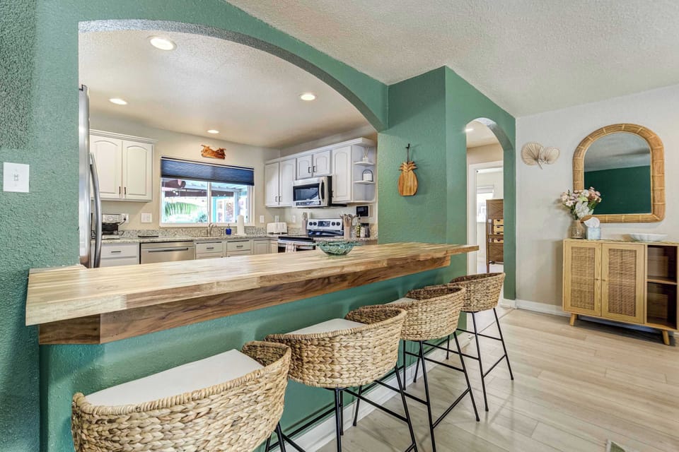 A wide arched opening frames the kitchen, where a wood bar top with three woven rattan barstools bridges the two rooms. White cabinetry and stainless appliances sit just beyond, keeping the cook in on the conversation.