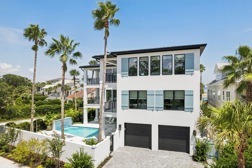Modern coastal architecture surrounded by lush tropical palms. The property features a private pool and spacious layout, nestled in a serene neighborhood with neighboring homes visible beyond the landscaped grounds.