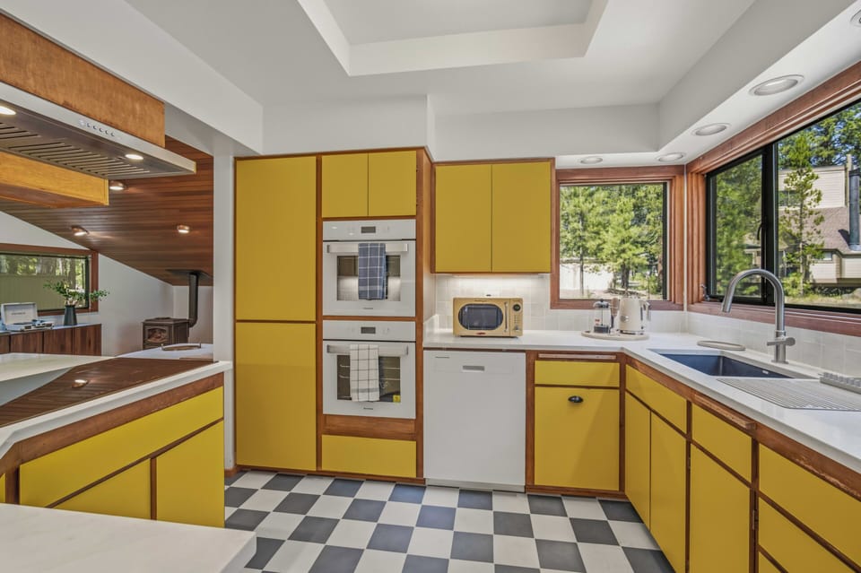 Step into your fully-equipped kitchen featuring cheerful retro-style cabinetry, modern appliances, and sunlit views of the surrounding trees through expansive windows—perfectly designed for creating memorable vacation meals together.