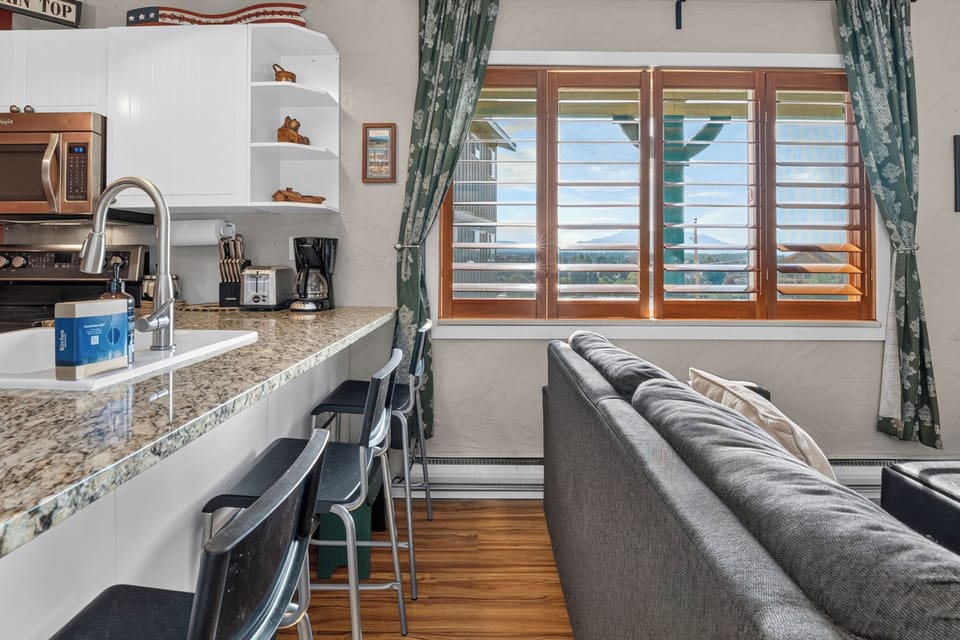 Your open-concept space blends kitchen and living area, with granite countertops, modern appliances, and a cozy sofa. Wooden shutters frame the windows, letting in warm natural light.