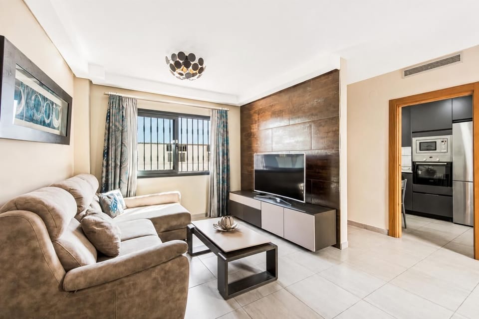 A contemporary living room features a plush brown sectional sofa, a textured accent wall with a wall-mounted TV, and an open doorway leading into a modern kitchen.