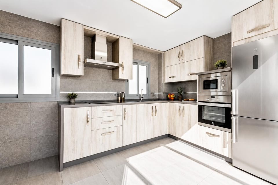 A modern kitchen features light wood grain cabinetry, gray tiled walls, and integrated stainless steel appliances in a bright, corner layout.