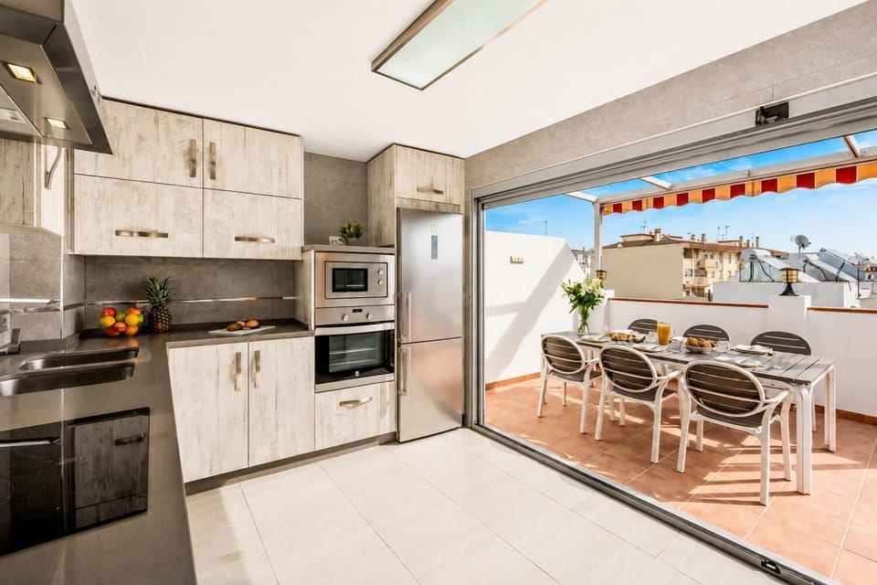 A modern kitchen with light-wood grain cabinetry opens through large glass doors directly onto a sunny rooftop terrace with an outdoor dining table.