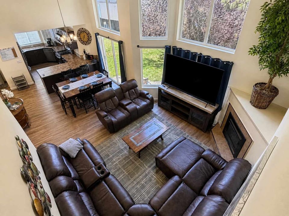 Elevated view of the open living, dining, and kitchen layout, featuring leather recliners, a large TV, fireplace, and tall windows.