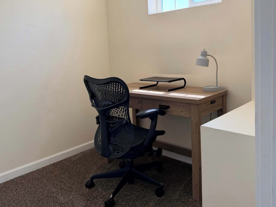 Dedicated work nook with a desk, adjustable office chair, task lamp, laptop stand, power access, and natural light.