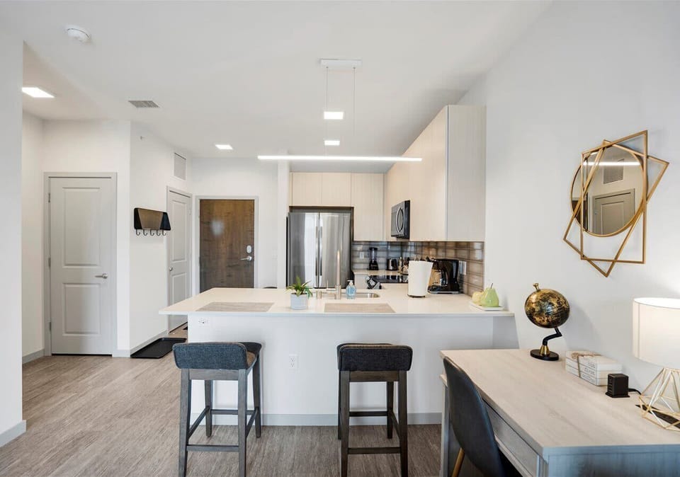 Bright open kitchen with sleek island seating for two, stainless steel appliances, and modern lighting. A stylish mirror and workspace add charm and functionality, perfect for cooking, dining, or working from home.