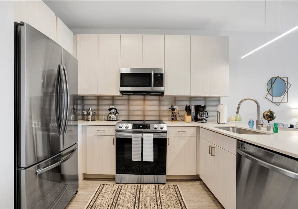Bright, modern kitchen with stainless steel appliances, sleek cabinetry, and a spacious island. Features a built-in microwave, full-size fridge, dishwasher, and elegant backsplash for a clean contemporary look.