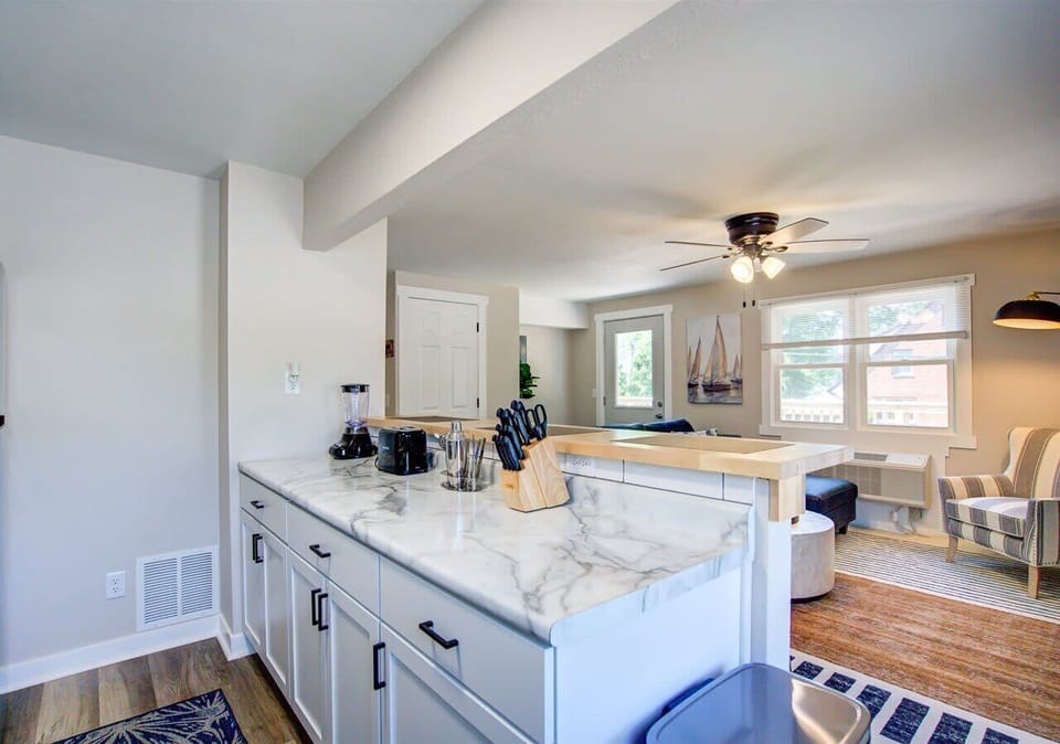 Bright open kitchen with marble-style counters, modern white cabinetry, and full stainless appliances. A raised breakfast bar with wood accents and seating overlooks the airy living space, creating a welcoming flow for cooking and casual dining.