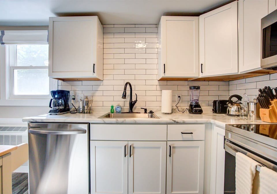 Bright, well-equipped kitchen featuring white cabinetry, sleek subway tile backsplash, and stainless steel appliances. Includes coffee maker, blender, and all the essentials for effortless meal preparation.
