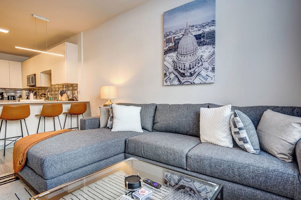 Stylish open-concept living room with a plush gray sectional, accent pillows, and a cozy throw. A sleek glass coffee table faces the kitchen with bar seating, creating a welcoming space to relax or entertain.