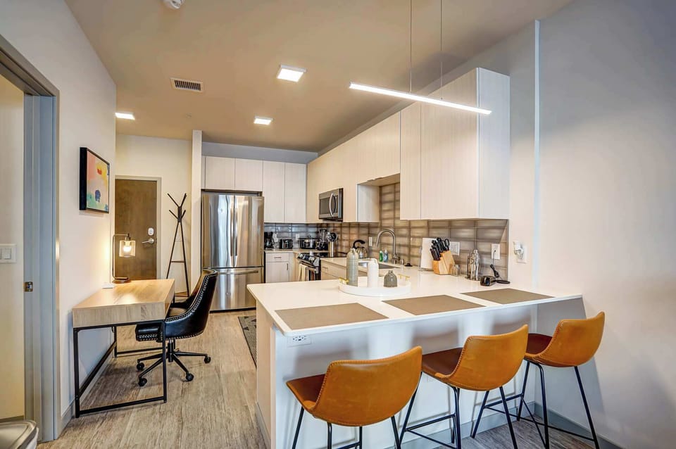 Open kitchen with sleek cabinets, stainless appliances, and a spacious island with three leather stools. A built-in desk and chair create a handy workspace near the entry, blending style and function in one inviting space.