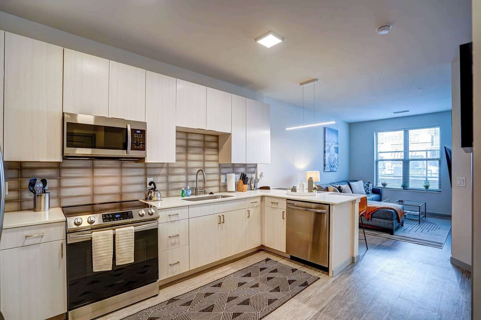 Open-concept kitchen with stainless appliances, sleek cabinetry, and a spacious island flows into a cozy living area with a large window for natural light—perfect for cooking and relaxing.