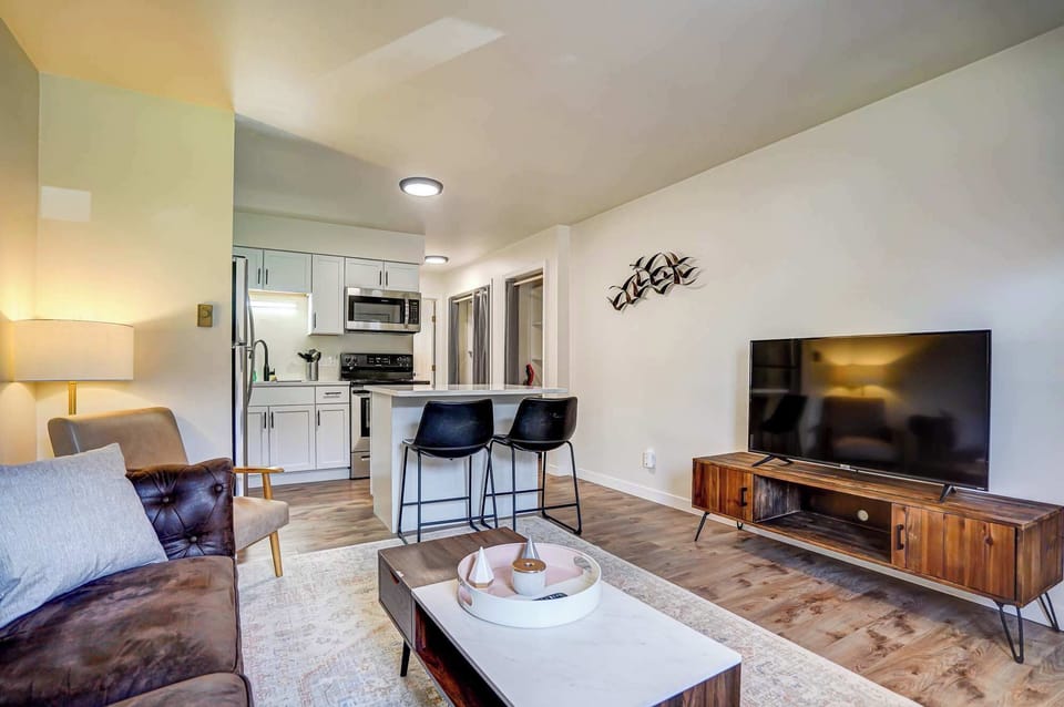 Bright and stylish open-concept living area featuring a cozy seating space, smart TV, and a modern kitchen with stainless steel appliances. The kitchen island doubles as a breakfast bar, perfect for casual dining or working from home.