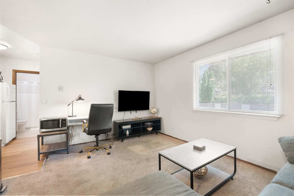 Bright and practical living space with a cozy sectional, dedicated work desk, and mounted 65" Roku Smart TV. The large window brings in natural light, creating an inviting atmosphere for relaxing or staying productive.
