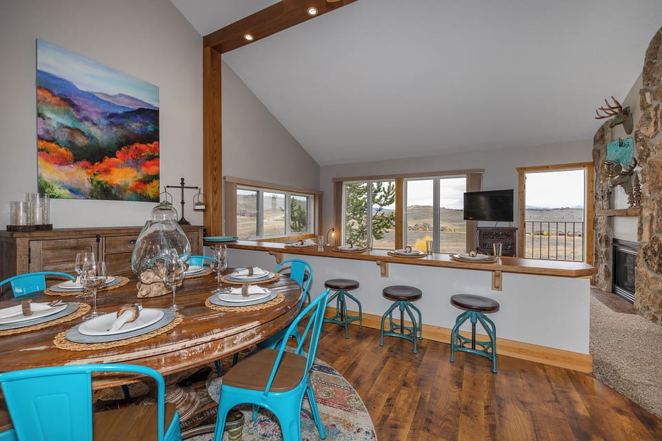 Step into your mountain retreat with a stunning open-concept dining and kitchen space featuring rustic wood beams, turquoise seating, and expansive windows framing sweeping views of the surrounding landscape.