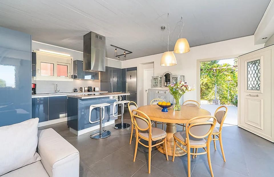 A modern open-plan kitchen and dining area features glossy blue cabinetry, a central island with bar stools, and a round wooden dining table under glowing pendant lights.