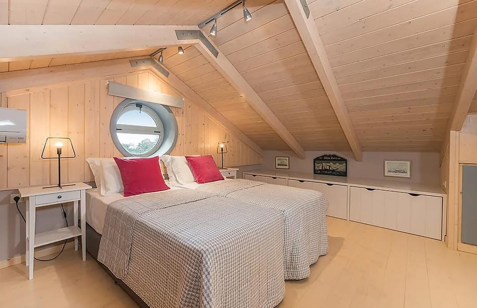 An attic bedroom features twin beds with patterned gray covers and bright magenta pillows beneath a sloped light wood paneled ceiling and a round porthole window.
