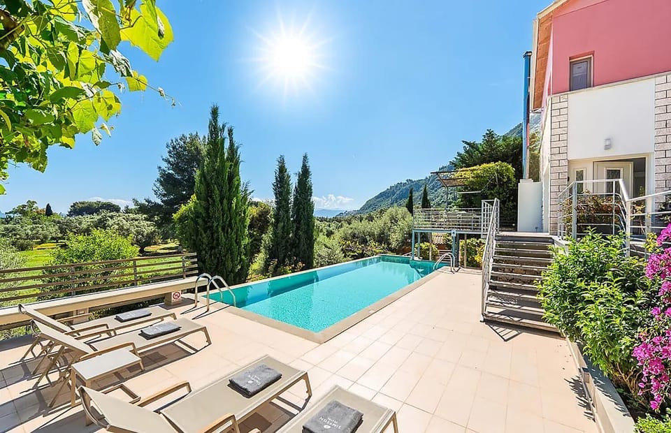 A sun-drenched outdoor swimming pool is lined with lounge chairs on a spacious tiled patio, offering a sweeping view of green trees and distant mountains under a bright blue sky.