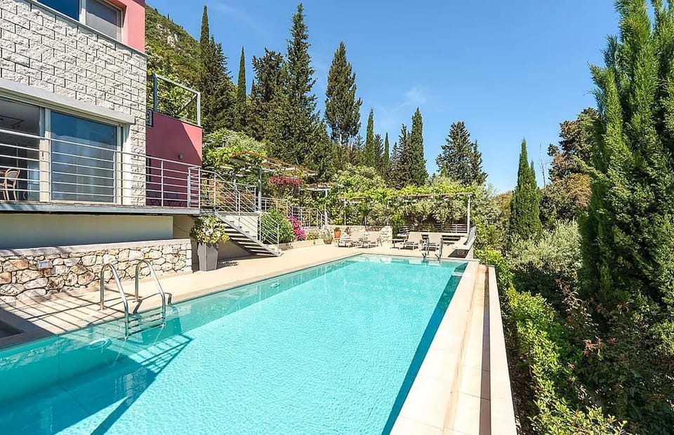 An outdoor swimming pool stretches alongside a multi-story stone and pink villa, surrounded by a spacious sun terrace and tall green cypress trees under a clear sky.