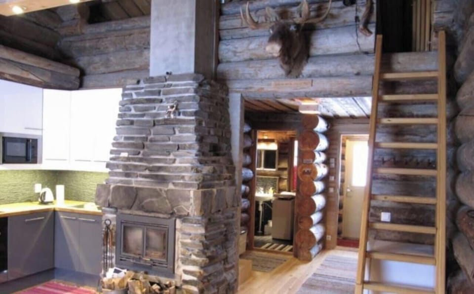 Log-walled living space with a stone fireplace, firewood at hand and a wooden stairway up to the loft.