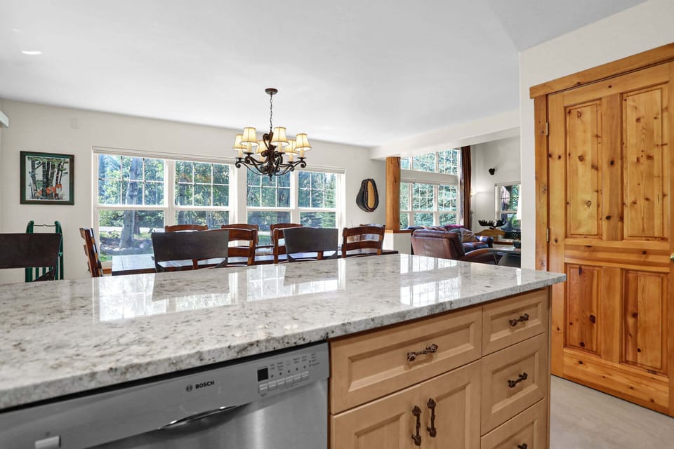 An open-concept kitchen island with speckled granite countertops and a stainless steel dishwasher looks out toward a spacious dining area illuminated by a classic chandelier and large windows.