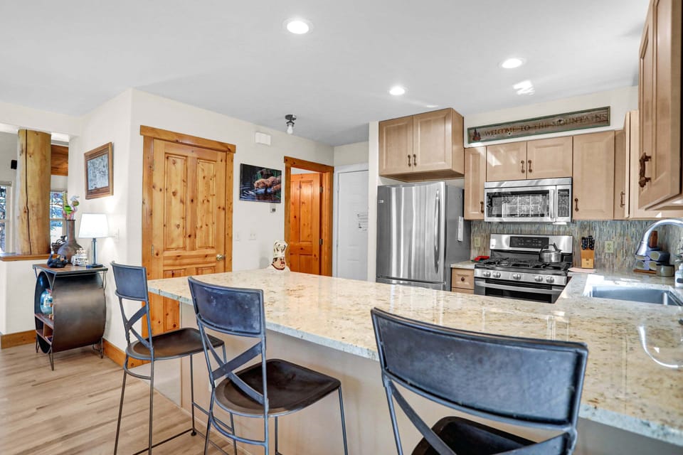 This warm, cabin-style kitchen features light wood cabinetry, speckled granite countertops, and stainless steel appliances alongside an expansive breakfast bar lined with modern barstools.