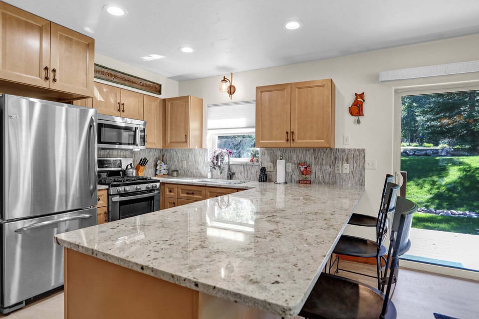 A bright, modern kitchen features light wood cabinetry, speckled granite countertops, and stainless steel appliances alongside a spacious breakfast bar looking out onto a lush green yard.