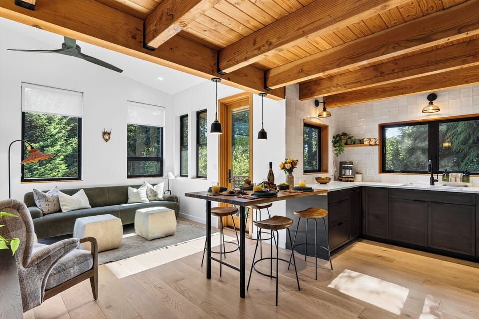 Airy kitchen and sitting area with exposed timber beams, breakfast bar seating, and forest-framed windows.