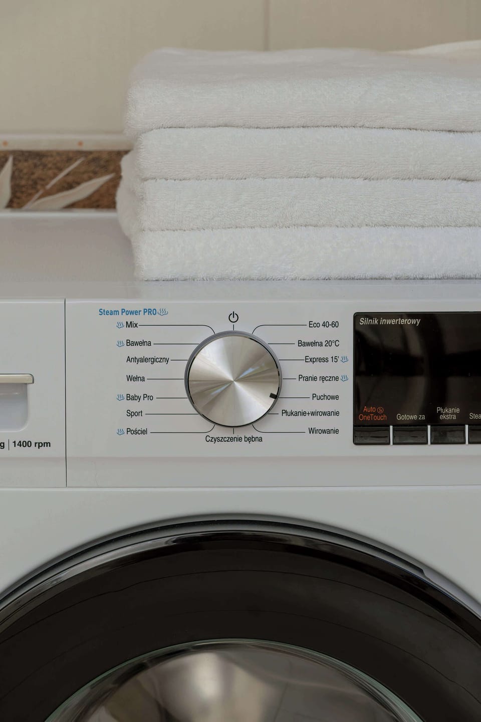 A close-up view highlights the control dial and display panel of a modern white washing machine with folded white towels resting on top. The focused detail emphasizes practical in-unit laundry amenities.