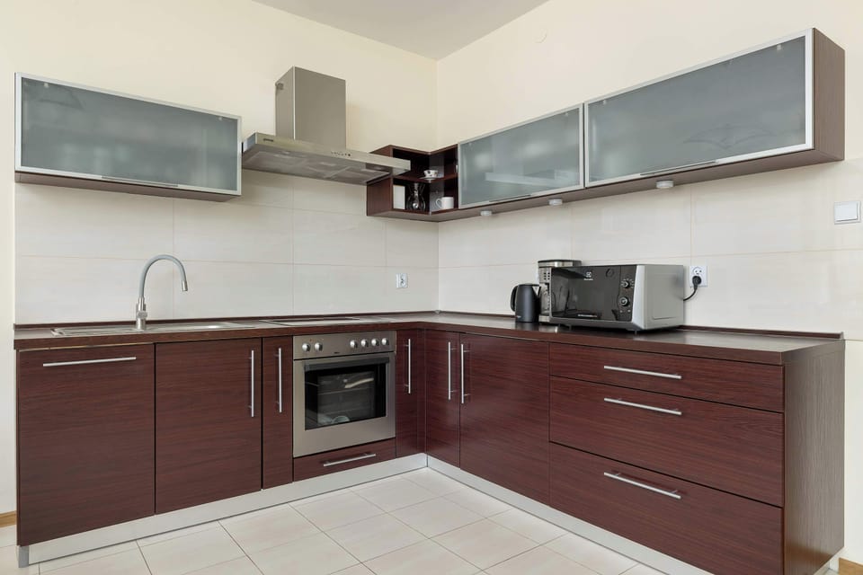 Dark wood lower and upper cabinets line a functional corner kitchen equipped with a built-in oven, stovetop, and countertop appliances. Light neutral tiles and walls keep the culinary area bright and tidy.